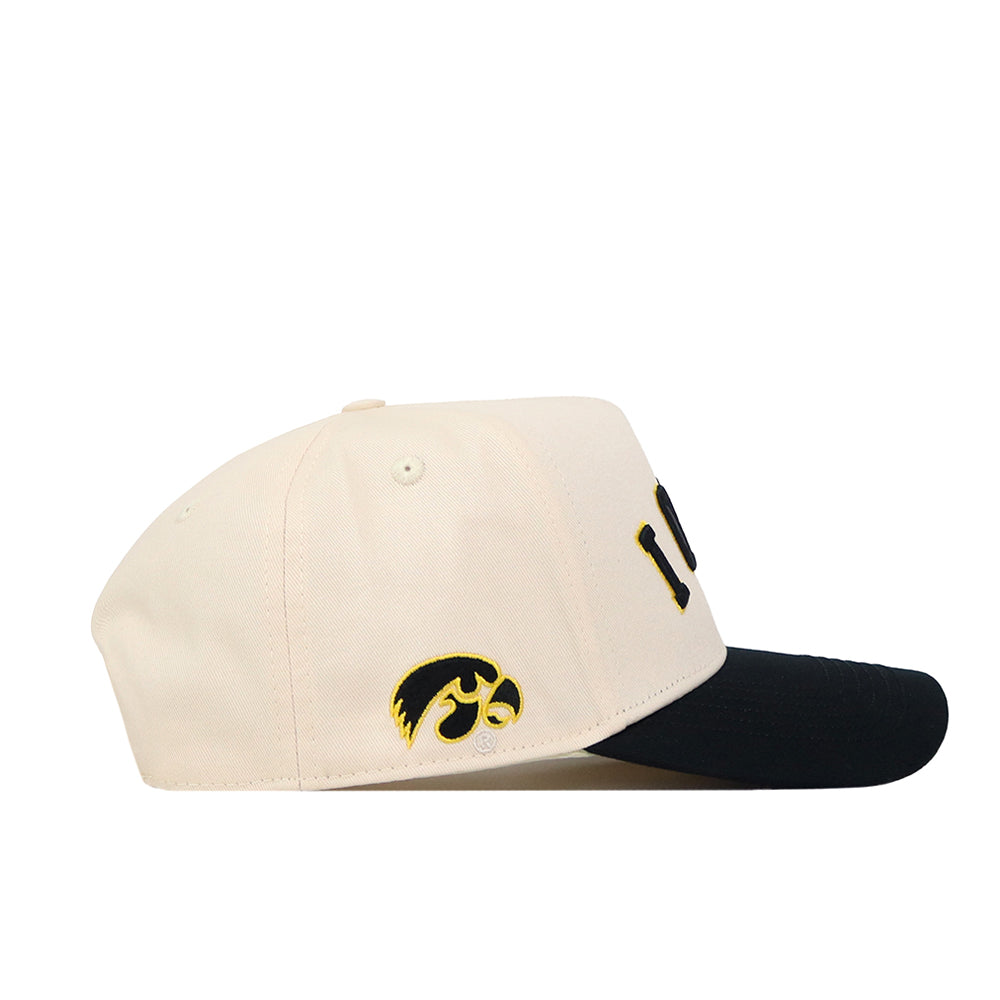 Side view of the No Rivals Right Side Up Iowa Hat – Classic, a cream-colored snapback with a black bill. The hat highlights an embroidered yellow and black Hawkeye logo on the side and partially visible black text on the front, making it the perfect finishing touch for your gameday look.