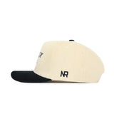 The Texas A&M Traditions Hat by No Rivals is a cream corduroy snapback with a black brim, shown from the side. The cap features embroidered NR initials in dark thread near the back and offers a structured, lasting fit.