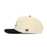 The No Rivals Alabama Traditions Hat is a cream snapback with a black brim, side view. Made of quality cotton, it has ventilation holes and “NR” embroidered in black near the back.