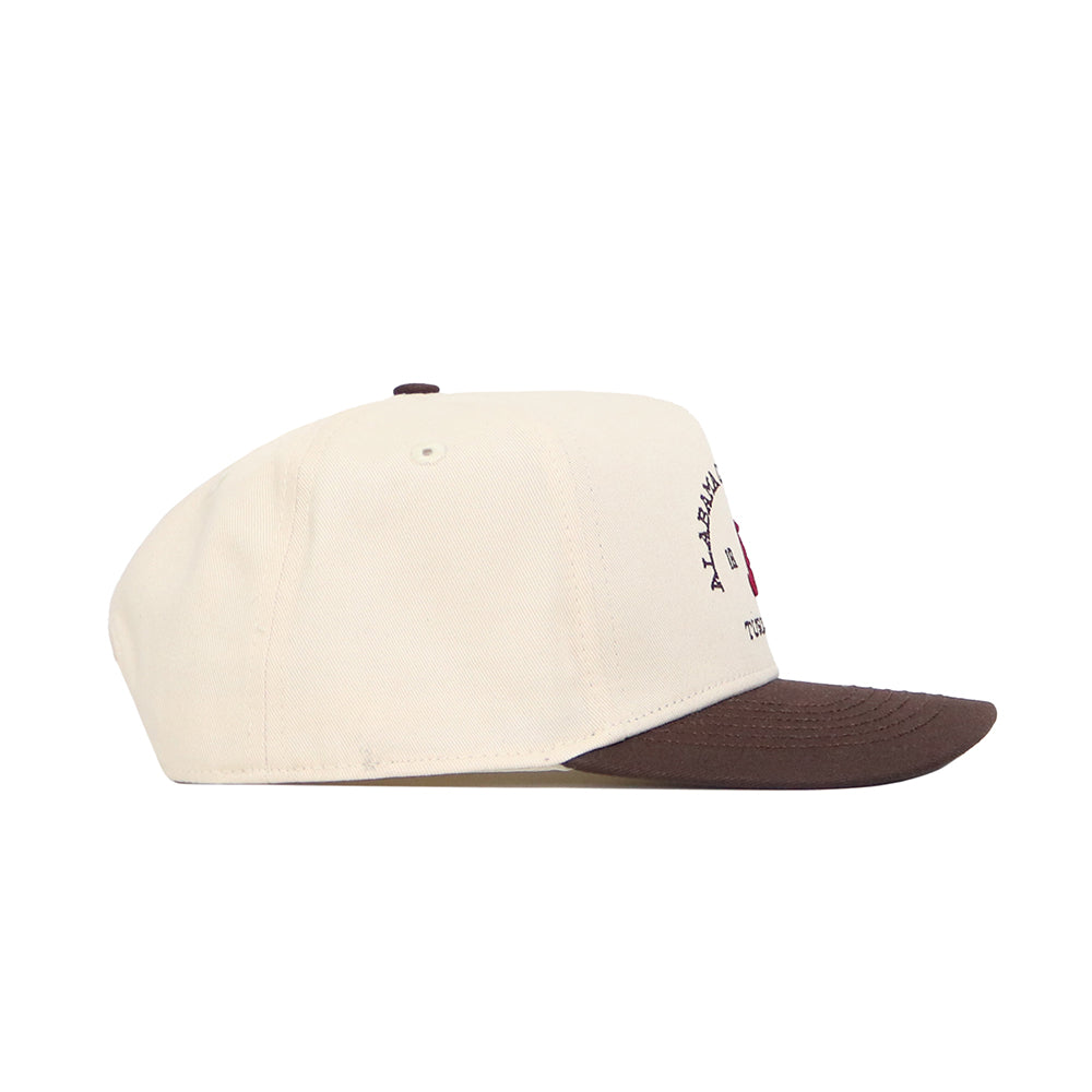 The No Rivals Alabama Outlaw Hat - Classic is a cream snapback with a brown brim and button, shown from the side. Made of quality cotton, it has front embroidery, though the text and details aren’t fully visible.