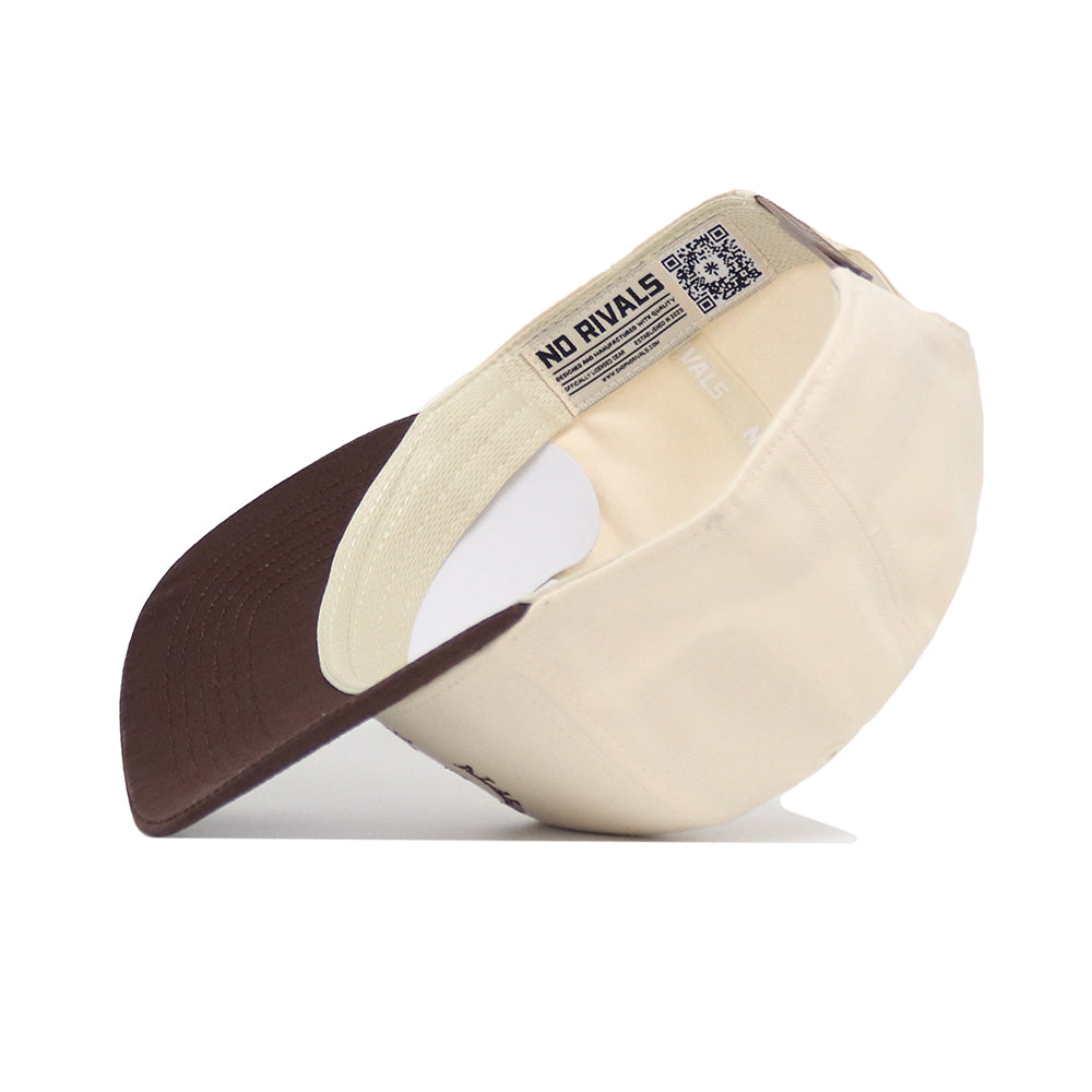The Alabama Outlaw Hat - Classic by No Rivals is shown upside down, revealing an inside label with the brand name and a QR code near the adjustable strap. The beige and brown cap features a structured fit and a pre-curved brown brim.