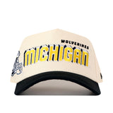 Introducing the Michigan Wolverines Alma Mater Hat by No Rivals—a cream and black baseball cap featuring an embroidered yellow and black MICHIGAN WOLVERINES logo on the front, a small football helmet graphic on the left side, and an adjustable snapback closure for a perfect gameday fit.