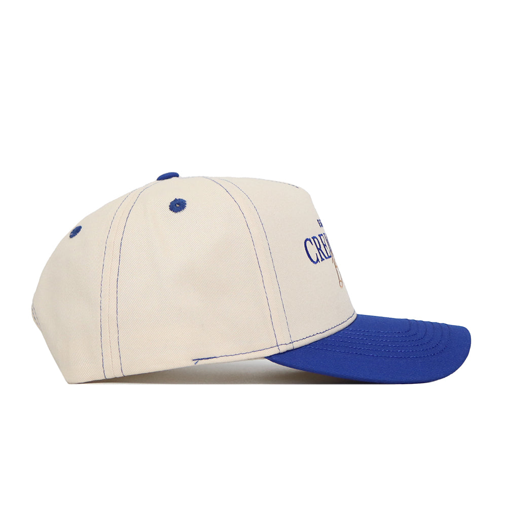 Side view of the Creighton Vintage Hat by No Rivals—a cream-colored baseball cap with blue stitching, a blue top button and curved brim, structured fit, and partially visible embroidered text on the front.