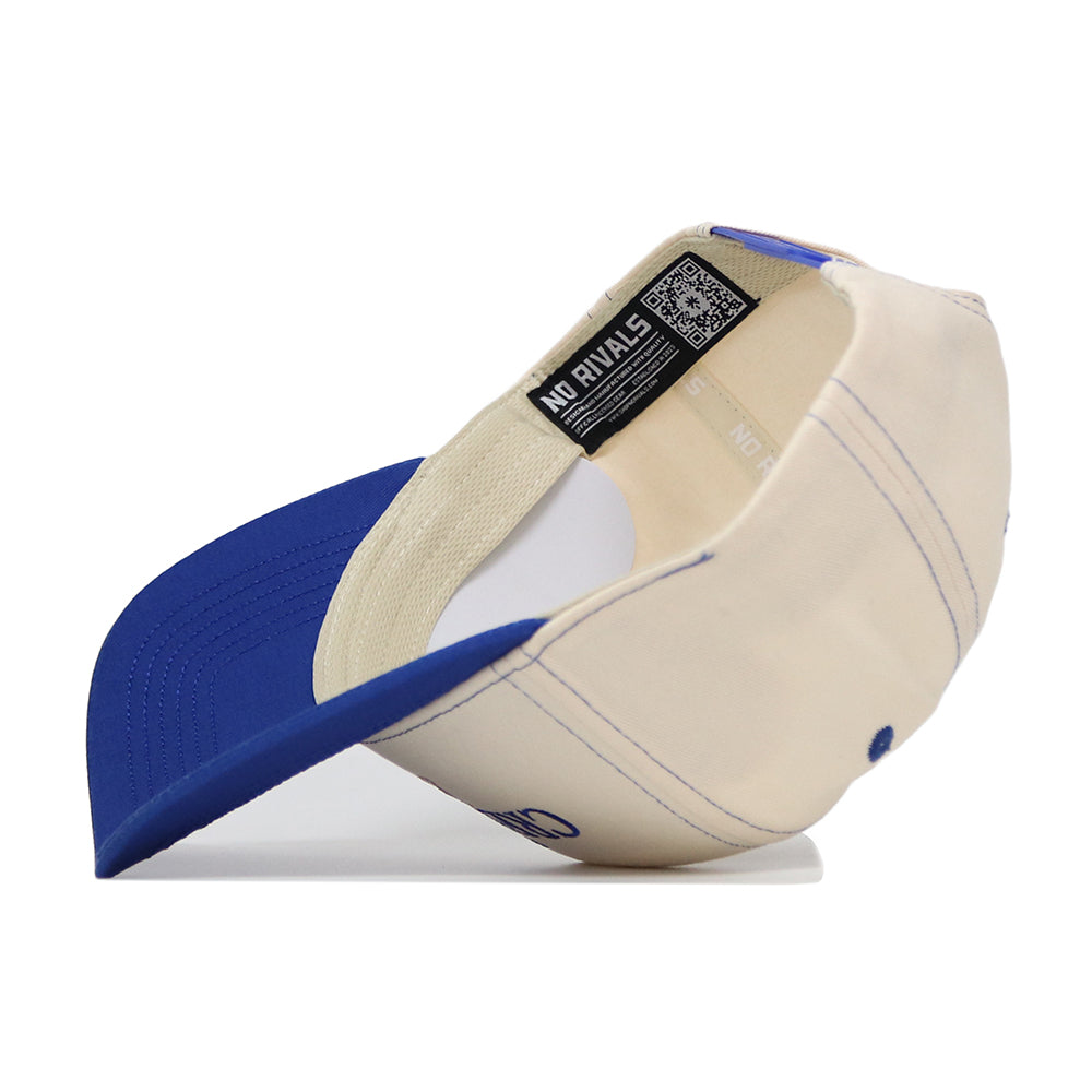 The Creighton Vintage Hat by No Rivals is shown upside down, displaying its interior label. This cream and blue baseball cap features a structured fit, visible stitching, and a curved blue brim.