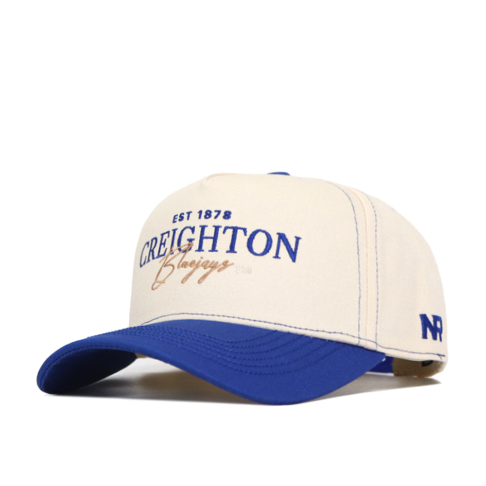 The Creighton Vintage Hat by No Rivals is a cream and blue baseball cap featuring "Est 1878 Creighton Bluejays" on the front, an "NR" logo on the side, structured fit, snapback closure, and official licensing. Bill and top button are blue.