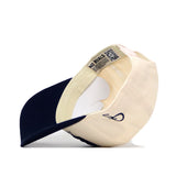 A beige and navy Georgia Southern Erk Russell Hat by No Rivals is pictured flipped upside down, revealing its interior lining, tags, and a circular cardboard insert that helps maintain the hat’s shape. The snap back closure offers an adjustable fit.