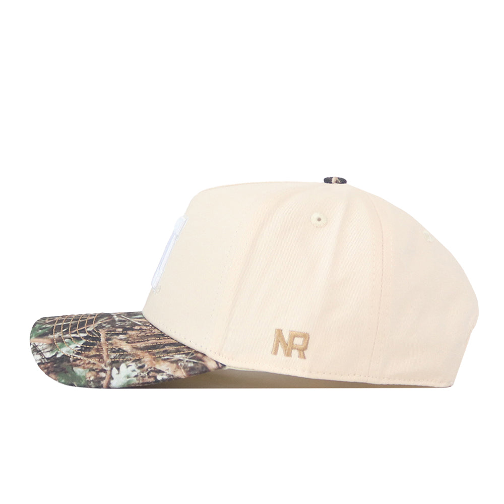 Introducing the Nebraska Camo Hat - Connector Edition by No Rivals: a beige baseball cap made from premium cotton, featuring a camouflage-patterned brim and gold “NR” initials embroidered on the side. This structured fit hat seamlessly blends comfort with standout style.