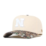 Introducing the Nebraska Camo Hat - Connector Edition by No Rivals: a beige cotton baseball cap featuring a white N logo on the front, complemented by a camouflage-patterned brim and a comfortable snap back closure, presented against a white background.