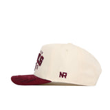 The Texas A&M Rally Hat by No Rivals is a cream snapback with a maroon brim, made from premium cotton. Bold maroon text decorates the front, with small NR initials embroidered in maroon on the side.