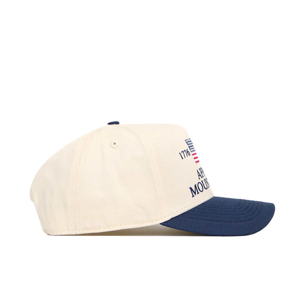 The No Rivals App State Mountaineers 250th Americana Hat features a cream crown, navy brim, visible stitching, a front embroidered logo, partial text, and an American flag graphic. The structured cap is shown in left profile.