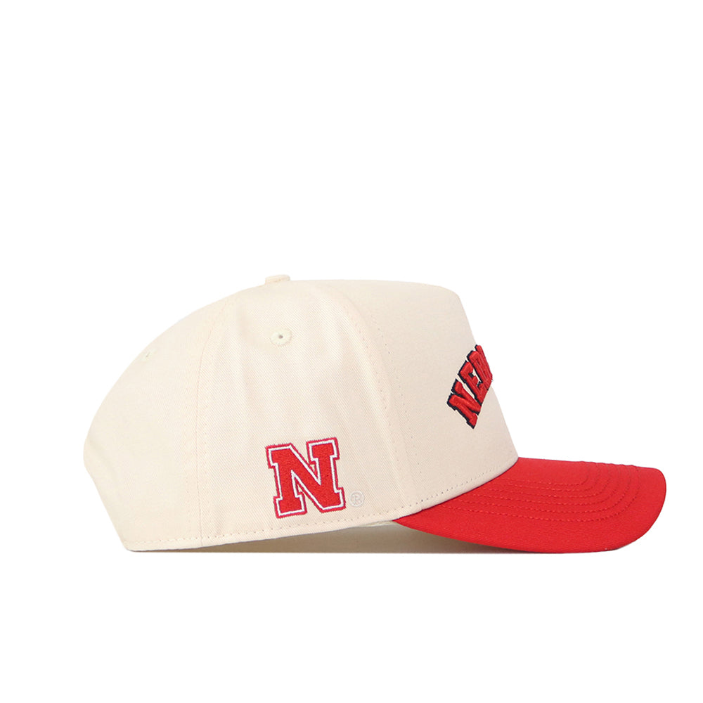 Introducing the Right Side Up Nebraska Hat - Classic by No Rivals: a cream-colored baseball cap crafted from high-quality cotton, featuring a striking red brim. This snapback hat is adorned with a bold red embroidered N logo on the side and eye-catching red and black partial lettering on the front, combining comfort and style for everyday wear.