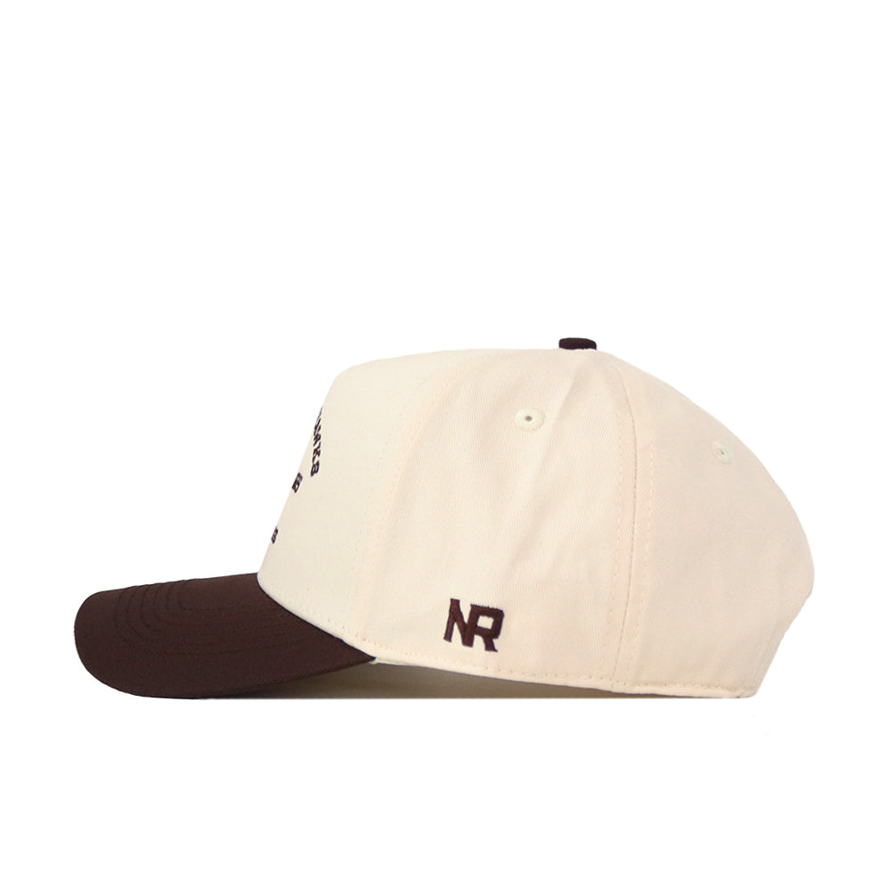 A side view of the Kansas Jayhawks Outlaw Hat - Classic by No Rivals, featuring a cream-colored crown and brown brim. Made from high-quality cotton, this cap showcases small brown embroidered NR initials near the back above the brim and offers a comfortable fit with its convenient snapback adjustment.