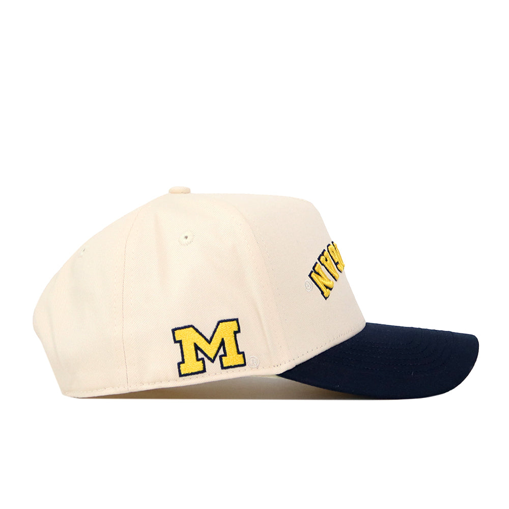 The Michigan Wolverines | Upside Down Michigan Hat - Classic by No Rivals is a beige baseball cap with a navy blue brim, showcasing a large yellow “M” on the side and bold yellow text across the front. Crafted from high-quality cotton, it features an embroidered logo for a premium finish.