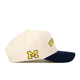 The Michigan Wolverines | Upside Down Michigan Hat - Classic by No Rivals is a beige baseball cap with a navy blue brim, showcasing a large yellow “M” on the side and bold yellow text across the front. Crafted from high-quality cotton, it features an embroidered logo for a premium finish.