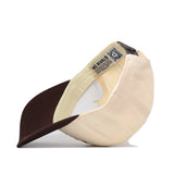 The Kansas Jayhawks Outlaw Hat - Classic by No Rivals is pictured here in a cream and dark brown color scheme, with its beige crown and deep brown bill. Shown flipped upside down, the image highlights the inside label, adjustable snapback strap for a comfortable fit, and a QR code tag on the inner band. Made from high quality cotton, this hat combines style and function for any fan.