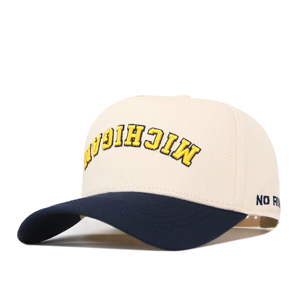 Introducing the Michigan Wolverines | Upside Down Michigan Hat - Classic by No Rivals—a beige and navy blue snapback cap made from premium cotton, showcasing “MICHIGAN” embroidered upside down in yellow across the front and a subtle “NO” logo on the side. This stylish cap blends comfort with a unique, eye-catching design.