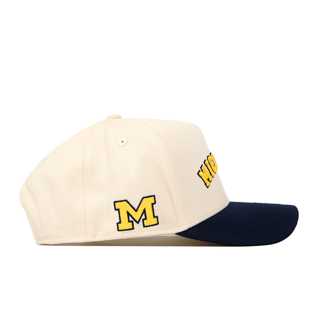 The Right Side Up Michigan Hat - Classic Colors by No Rivals is a cream snapback with a navy brim, featuring an officially licensed yellow M logo embroidered on the side and partial yellow text on the front.