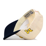 The Right Side Up Michigan Hat - Classic Colors by No Rivals is shown lying on its side, featuring a cream and navy design with a yellow embroidered letter W on the side and an inside tag that reads NO RIVALS.