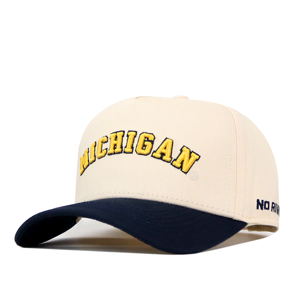 The Right Side Up Michigan Hat - Classic Colors by No Rivals features a beige snapback design with “MICHIGAN” embroidered in yellow letters on the front and a navy blue brim. The right side of the brim displays small navy text reading “NO RIVALS.” This officially licensed cap blends classic style with true team pride.
