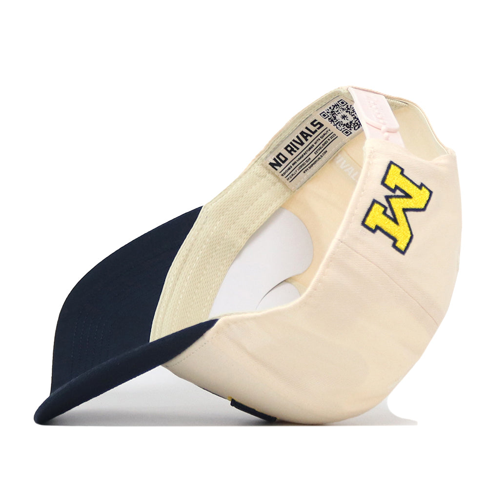 The Michigan Wolverines Stadium Hat by No Rivals, shown in beige and navy, is pictured flipped upside down to reveal an embroidered yellow M logo on the side and an interior label that reads “NO RIVALS.” This snapback cap features a curved bill, visible interior stitching, and offers a classic gameday fit.