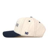 A cream and navy Michigan Wolverines Vintage Hat by No Rivals, shown from the side, featuring an officially licensed embroidered NR logo near the back, a navy bill, and a classic snap back closure.