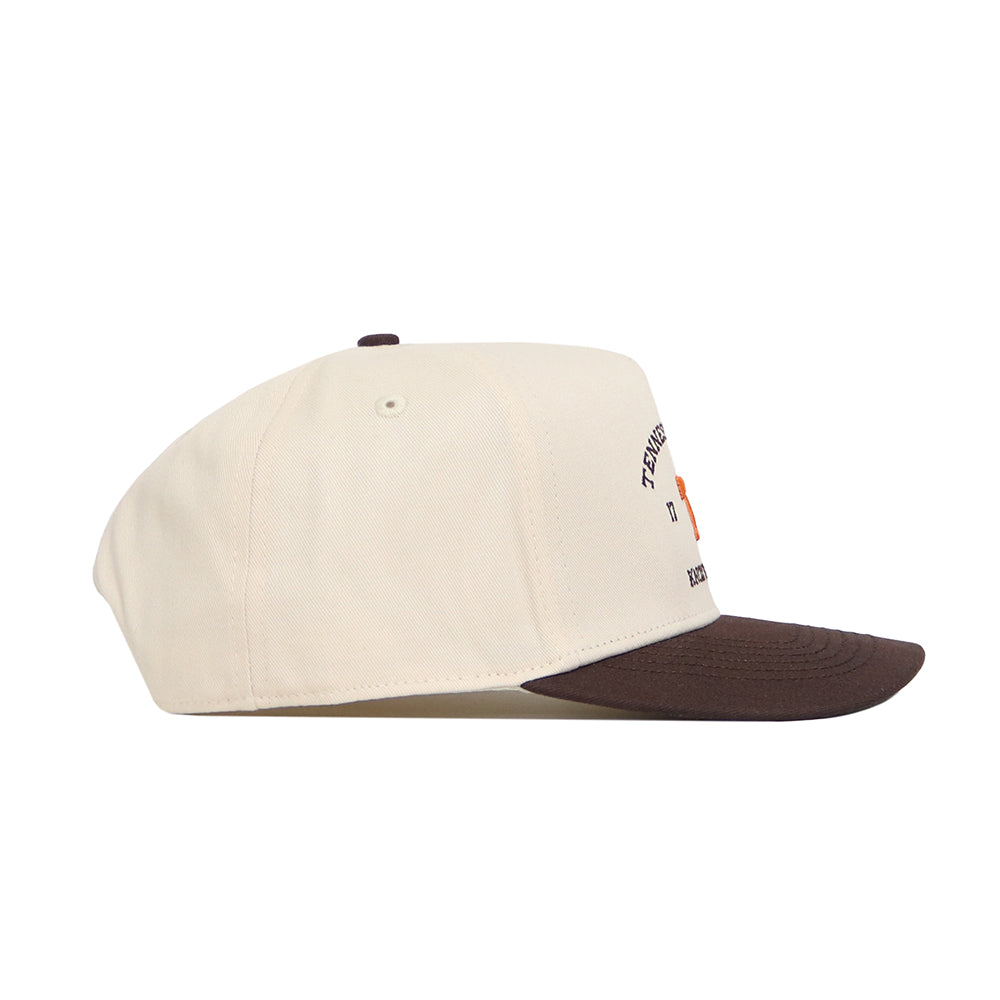 The Tennessee Outlaw Hat - Classic by No Rivals is a cream snapback with a dark brown brim, shown in a side view. Made from premium cotton, it displays partially visible small text and graphics on the front panel.