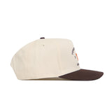 The Tennessee Outlaw Hat - Classic by No Rivals is a cream snapback with a dark brown brim, shown in a side view. Made from premium cotton, it displays partially visible small text and graphics on the front panel.