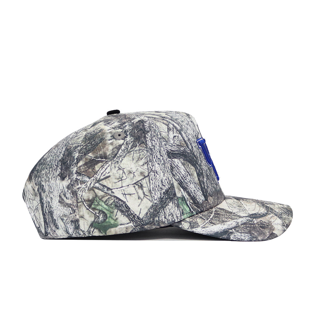 The Kentucky Outland Hat by No Rivals is a camouflage snapback with a flat brim, featuring gray, brown, and green branch and leaf patterns and a blue embroidered patch on the structured cotton front. Side view shown.