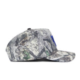 The Kentucky Outland Hat by No Rivals is a camouflage snapback with a flat brim, featuring gray, brown, and green branch and leaf patterns and a blue embroidered patch on the structured cotton front. Side view shown.