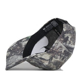 The Wichita State Outland Hat by No Rivals is shown tilted on its side, highlighting the interior lining and tags. Its snap back and forest camo bill in green, brown, and tan face forward.
