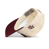The No Rivals Texas A&M Stadium Hat is shown from an angled side view, featuring a structured beige and maroon snapback design with ATM embroidered on the back, a maroon bill, and visible cotton inner tag with QR code.