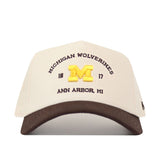 The No Rivals Michigan Wolverines Outlaw Hat - Classic showcases a cream and brown design with a prominent, embroidered yellow “M” logo on the front, along with “MICHIGAN WOLVERINES,” “18 17,” and “ANN ARBOR, MI” stitched detailing. Finished with a classic snap back closure for an ideal gameday fit.