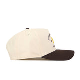 A cream-colored Michigan Wolverines Outlaw Hat - Classic by No Rivals, featuring a dark brown brim and partially visible embroidered logo and text on the front panel, is shown from the side. This stylish snapback is perfect for your next gameday fit.