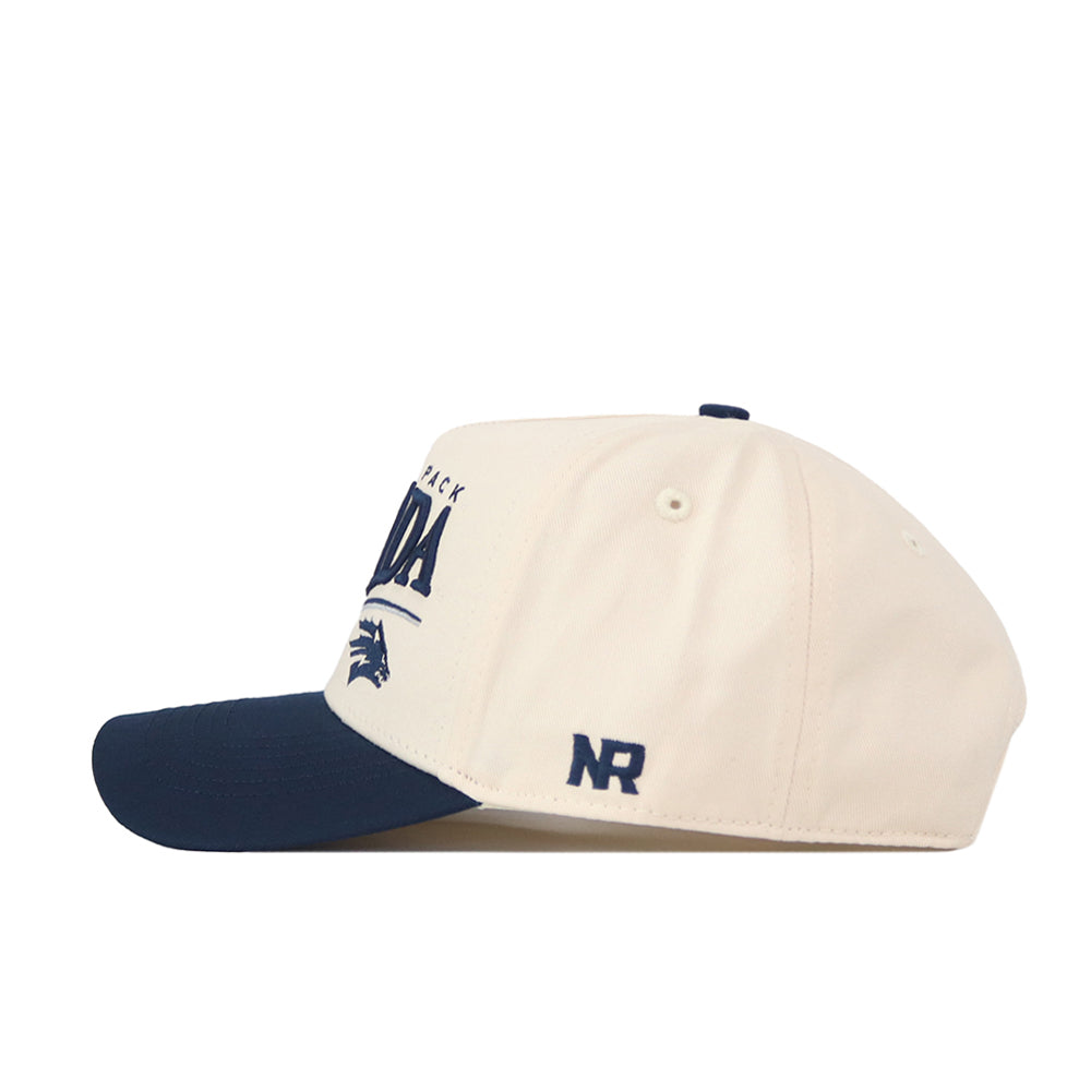 Introducing the Nevada Wolf Pack Stride Hat by No Rivals—a cream-colored baseball cap with a navy blue brim, expertly crafted from high-quality cotton. This stylish snapback features embroidered Nevada text, a distinctive wolf logo on the side, and the initials “NR” near the back for added flair. Combining standout details with everyday comfort, it’s the perfect accessory for showcasing your Wolf Pack pride.
