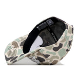 The Sam Houston State Huntsman Hat by No Rivals is displayed upside down, revealing the interior with a black sweatband bearing the NO RIVALS label. This snap back cap features a green, beige, and black camouflage pattern and a flat bill. Made from high-quality cotton, it also showcases an embroidered logo for enhanced style and durability.