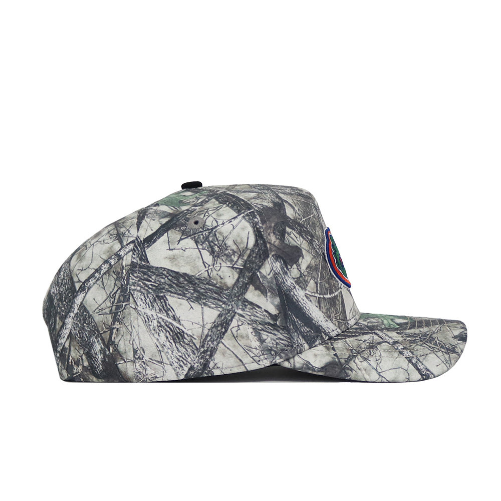 Side view of the No Rivals University of Florida Outland Hat, a structured fit snapback with a camouflage pattern and multicolored circular logo on the front; background shows tree branches and leaves.