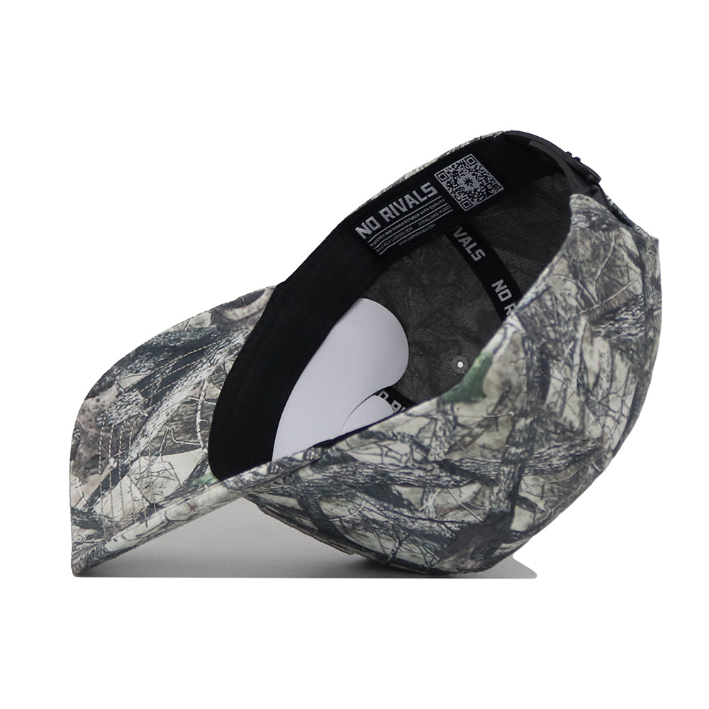 The Hawaii Outland Hat by No Rivals is shown upside down on a white surface, revealing the inside label. This snapback cap features a camouflage pattern in grey, green, and black and is made from high-quality cotton.