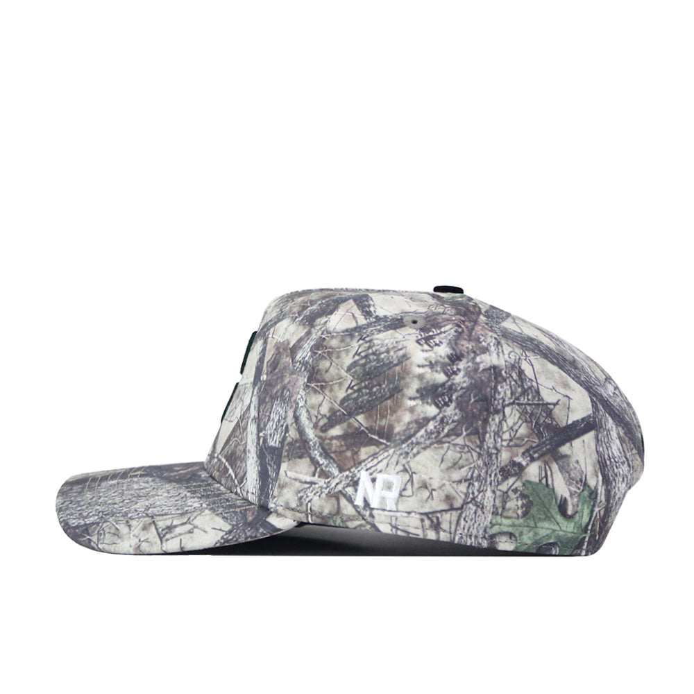 The Charlotte Outland Hat by No Rivals is a camouflage baseball cap with a flat brim, shown in profile facing left. Its earthy-toned pattern features branches and leaves, while the side displays a bold, white embroidered “NP” logo. The hat includes a convenient snapback adjustment for the perfect gameday fit.
