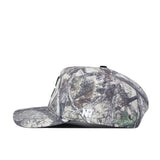 The Charlotte Outland Hat by No Rivals is a camouflage baseball cap with a flat brim, shown in profile facing left. Its earthy-toned pattern features branches and leaves, while the side displays a bold, white embroidered “NP” logo. The hat includes a convenient snapback adjustment for the perfect gameday fit.
