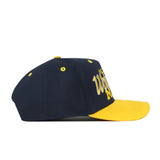 The No Rivals Michigan Wolverines Boomerang Hat is a navy blue snapback baseball cap with a yellow brim and a yellow embroidered logo on the front, shown from the side. Yellow eyelets are visible on the crown.