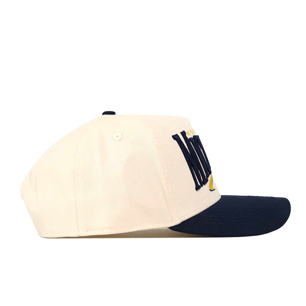 The No Rivals Michigan Wolverines Stride Hat features a cream-colored snapback design with a navy blue brim and raised embroidered lettering on the side. This officially licensed cap is displayed from a side angle against a white background.