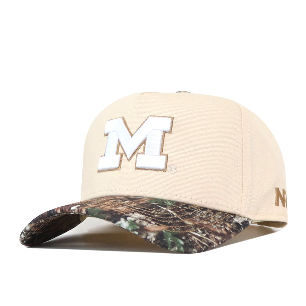 The No Rivals Michigan Wolverines Camo Connector Hat is a beige snapback baseball cap featuring a large white embroidered ‘M’ on the front, a camouflage-patterned brim, and small gold text detailing on the side.