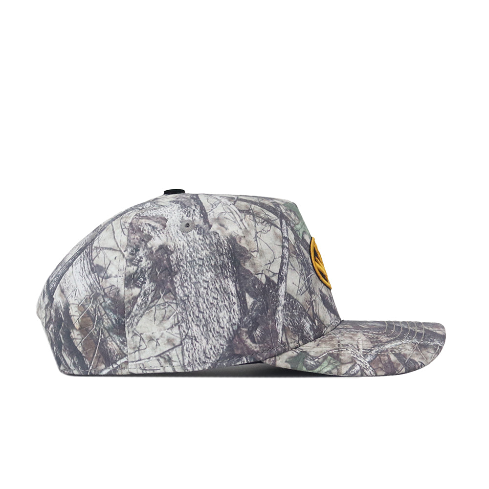 Side view of the No Rivals Mizzou Outland Hat, a camouflage baseball cap featuring a yellow patch on the front. This hat showcases a blend of green, brown, and gray natural patterns that resemble leaves and branches.