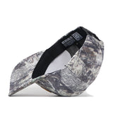 The No Rivals Mizzou Outland Hat is shown on its side, revealing the underside of the bill and the inside label with text and a QR code. This camouflage baseball cap features a structured fit for comfortable wear, combining style and practicality.