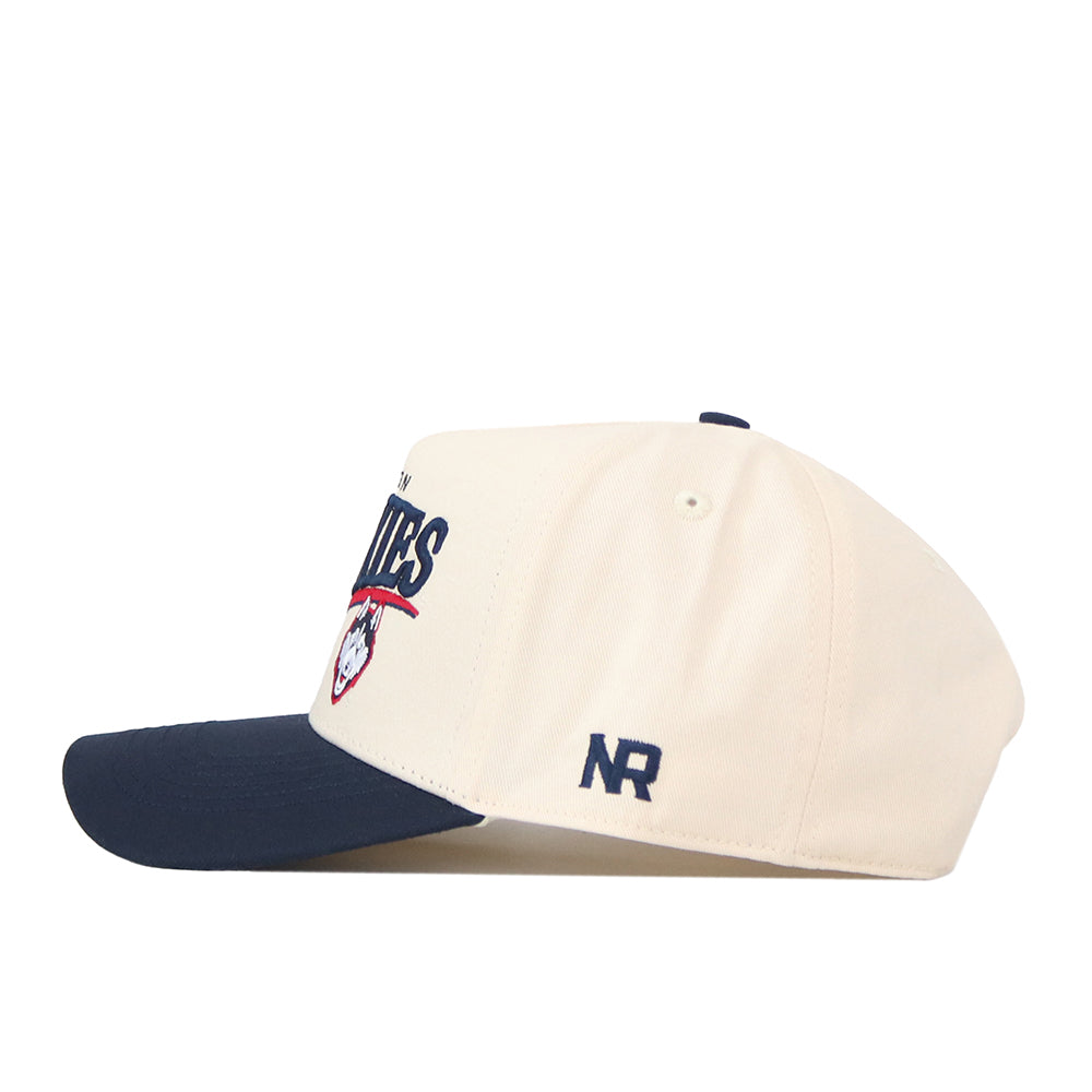 The UConn Huskies Stride Hat by No Rivals is a cream-colored baseball cap with a navy blue brim, crafted from high-quality cotton. It features the UConn Huskies sports logo on the side and the initials “NR” embroidered above the ear area. This structured fit snapback offers both comfort and style for everyday wear.