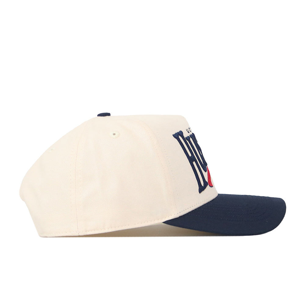 The No Rivals UConn Huskies Stride Hat is a cream-colored baseball cap with a navy blue brim, displayed in side profile. Its front showcases a bold, dark graphic with striking red accents. Made from high-quality cotton, this structured-fit snapback hat blends everyday comfort and style.