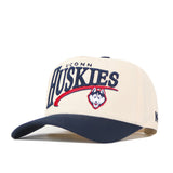 The No Rivals UConn Huskies Stride Hat is a structured fit cap in cream and navy, featuring embroidered UConn Huskies text and a husky mascot logo on the front. Crafted from high-quality cotton, it has an adjustable snap back closure for a custom fit.