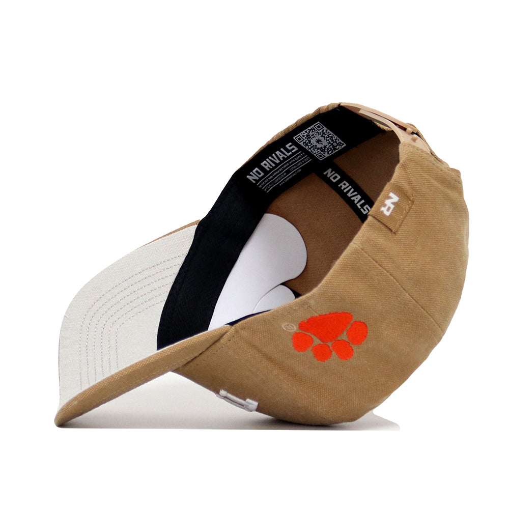 The Clemson Rustic Hat by No Rivals is a tan cotton canvas cap with an orange paw print logo on the side. When flipped, it reveals a white inner lining and black sweatband featuring the text "NO RIVALS" inside.