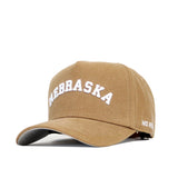 The Nebraska Rustic Hat by No Rivals is a tan cotton canvas baseball cap with "NEBRASKA" embroidered in white on the front and "NO RISK" on the brim side. It features a snap back closure for adjustable fit.