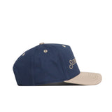 The No Rivals Sunday Golf Hat in navy and tan features a beige brim and button, a side-view white embroidered logo on the front panel, metal eyelets for ventilation, and adds style to your gameday fit.